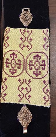A black piece of cloth with gold colored clasps between the two clasps is a piece of handwoven samite witha yellow background, purple ivy in columns and a purple circle enclosing a fluery cross.
