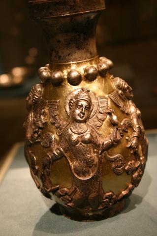 A silver vase depicting a women wearing a robe with a thin piece of cloth draped between her hands.